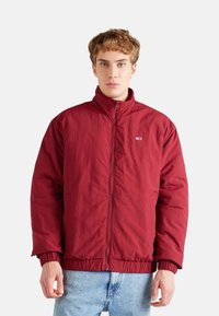 Chaqueta aislante roja con cuello alto, cierre de cremallera, puños y dobladillo elásticos. Presenta un parche con el logo en el pecho. La lleva puesta sobre unos vaqueros de color azul claro.