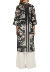 Schwarzer transparenter langer Mantel mit floralen Mustern in Creme, mit Dreiviertelärmeln und geradem Schnitt, getragen über weißen weit geschnittenen Hosen.