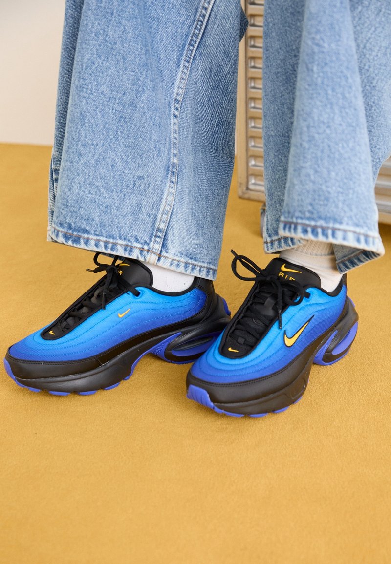Feet wearing blue and black Nike Air sneakers with yellow swoosh logos, white socks, and wide-leg blue jeans on a yellow floor.