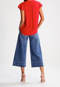 Blouse rouge à manches courtes avec un ourlet arrondi, associée à un jean en denim bleu à jambes larges. Présente des ourlets effilochés et un lavage clair. Portée avec des talons roses.