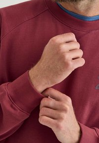 Felpa bordeaux con collo a girocollo e polsini a costine. Dettagli di cucitura visibili e una superficie testurizzata vicino al polso.