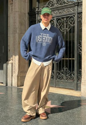 Jonge man met een groene pet, blauwe trui met "THE LAST EDITION," wit shirt, wijde beige broek en bruine schoenen, zittend voor een versierd ijzeren hek.