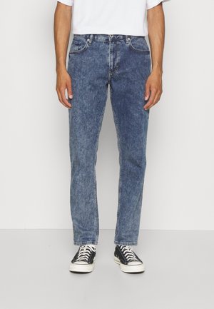 Mand iført syrevaskede blå jeans, sorte og hvide sneakers og en hvid t-shirt, stående mod en ensfarvet lys baggrund.