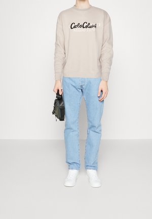 Felpa beige con testo del logo bianco e nero, jeans azzurri chiari e sneakers bianche. Tiene una piccola borsa nera con tracolla.