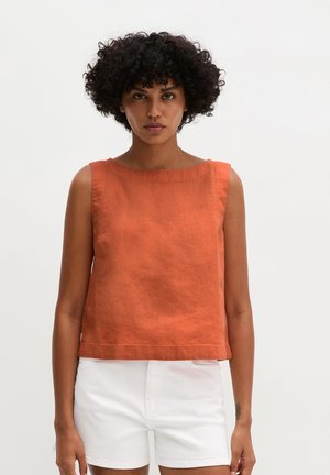 Frau mit lockigem Haar, die ein ärmelloses oranges Top und weiße, hoch taillierte Shorts trägt, steht vor einem schlichten hellen Hintergrund.