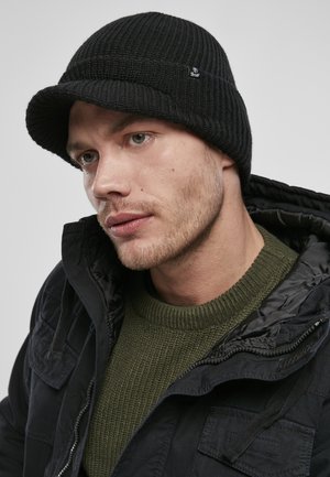 Must hägune soonikkohta beanie, millel on äär; kantud tumeda jope ja tekstuuriga oliivrohelise kampsuniga. Beanie'l on märkimisväärne brändimine.