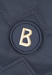 Tessuto trapuntato di colore blu navy con cuciture a zigzag; emblema rotondo con una lettera 'B' dorata centrata su uno sfondo nero.