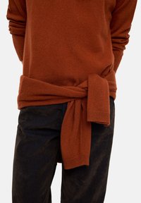 Sweter w kolorze rdzy z wcięciem w pasie, zestawiony z ciemnymi, teksturowanymi spodniami. Materiał wydaje się miękki i przytulny.