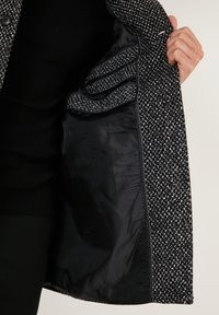 Abrigo de tweed en blanco y negro, con un exterior texturizado, forro negro suave y tres bolsillos interiores; detalles de botones visibles.
