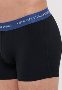 Fekete boxer alsónadrág kék elasztikus derékpánttal, amelyen fehér szöveggel szerepel a "Copenhagen Studios" felirat, kellemesen testhezálló fazonnal.