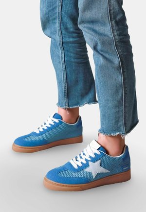 Personne portant des baskets en mesh bleu avec une étoile blanche et des lacets blancs, associées à un jean bleu raccourci à ourlet effiloché.