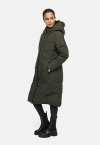 Manteau long matelassé vert olive avec capuche, présentant un design matelassé, une fermeture éclair à l'avant et un patch de logo. Associé à des bottines noires et un jean.