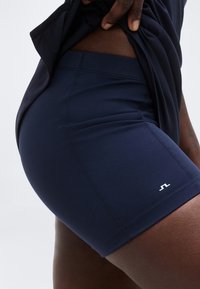 Záber zblízka na osobu, ktorá si ťahá tmavomodré športové šortky s malým bielym logom na stehne na jednoduchom svetlom pozadí.