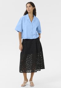 Camicia azzurra cropped con bottoni e orlo arricciato, abbinata a una gonna midi nera in pizzo con motivo geometrico e design a rete aperta.