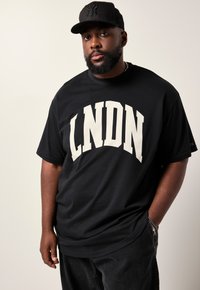 Sort t-shirt med hvid "LNDN" tekst over forsiden; parret med mørke bukser og en sort baseballkasket med logo.