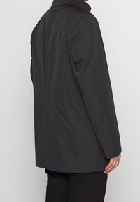 Czarna wodoodporna kurtka o prostym fasonie, gładkiej fakturze, bocznych kieszeniach i mankiecie zapinanym na guziki. Widok od tyłu.