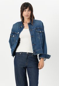 Mørkeblå denimjakke med knappelukking foran, to brystlommer og rullede ermer, kombinert med en hvit tank topp og mørke jeans.
