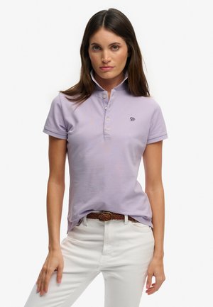 Kobieta ubrana w jasnofioletową koszulkę polo z krótkim rękawem i białymi guzikami, białe spodnie oraz brązowy pleciony pasek, stojąca na jednolitym tle.