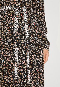Robe noire à fleurs avec un motif vibrant de fleurs, une fermeture à boutons et une ceinture avec la marque "TOMMY JEANS" en blanc.
