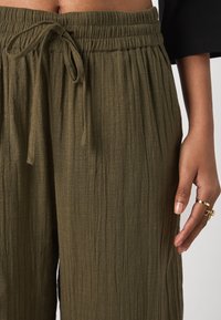 Pantalon léger vert olive texturé avec une taille élastique et un cordon de serrage, doté d'une coupe décontractée et de plis verticaux.