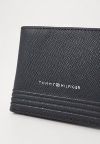 Zwarte gestructureerde portemonnee met reliëfontwerp, voorzien van een wit logo "TOMMY HILFIGER" en horizontale lijndetails nabij de basis.