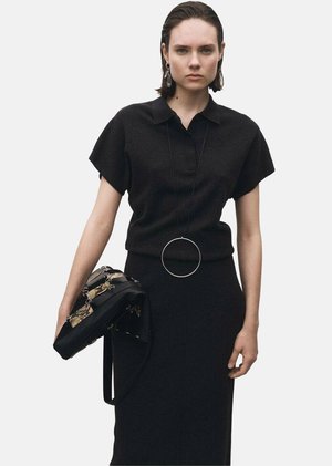 Haut en maille noire à manches courtes, associé à une jupe noire ajustée. Complété par un collier avec pendentif circulaire et un sac à main noir avec des accents dorés.