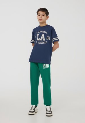 Granatowa bawełniana koszulka z białym nadrukiem 'LA' i pasami na rękawach, zestawiona z zielonymi spodniami dresowymi z napisem '1986'. Uzupełniona czarno-białymi trampkami.