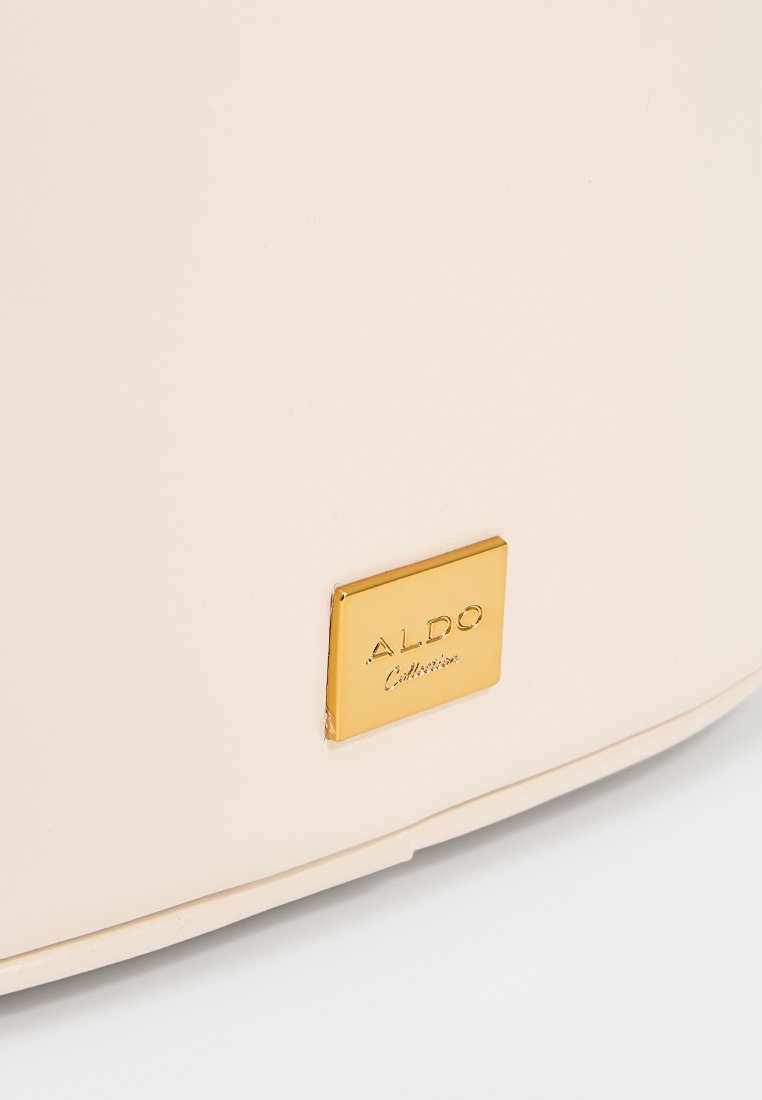 Helle beige Handtasche mit glatter Textur, ausgestattet mit einer goldenen quadratischen Logo-Plakette, die auf der Vorderseite mit "ALDO Collections" geprägt ist.