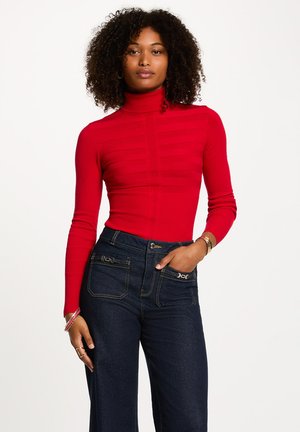 Maglione rosso a collo alto a coste con maniche lunghe, caratterizzato da una texture a strisce sul petto, abbinato a jeans in denim scuri a vita alta.
