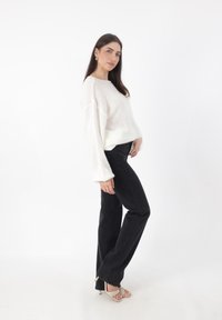 Witte gebreide trui met brede mouwen, gecombineerd met zwarte flare jeans en beige sleehakken. Model in een zijaanzicht pose.