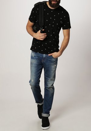 Mann trägt schwarzes T-Shirt mit Aufdruck, blaue zerrissene Jeans, schwarze Turnschuhe, trägt einen schwarzen Rucksack über einer Schulter und geht vorwärts.