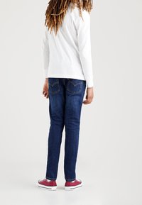 Vista trasera de una persona que lleva jeans de mezclilla azul oscuro, una camisa blanca de manga larga y zapatillas rojas con suelas blancas. Diseño notable del bolsillo trasero.