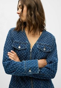 Chaqueta de denim con cierre de cremallera en la parte delantera, escote en V y dos bolsillos en el pecho. Presenta un patrón azul texturizado, costuras en contraste y acentos en plata.