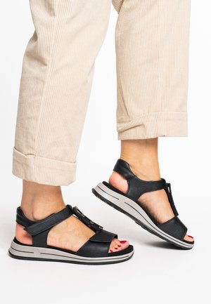 Schwarze Ledersandalen mit T-Riemen-Design, ausgestattet mit einer silbernen Schnalle, auf einer gepolsterten grauen Sohle; zeigen offene Zehen- und Fersenausschnitte.