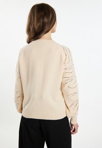 Beige svetr s dlouhými, měkkými rukávy, na kterých jsou kulaté flitry v vlnitých vzorech. Hladký límec a lem, volný střih, pletená struktura.