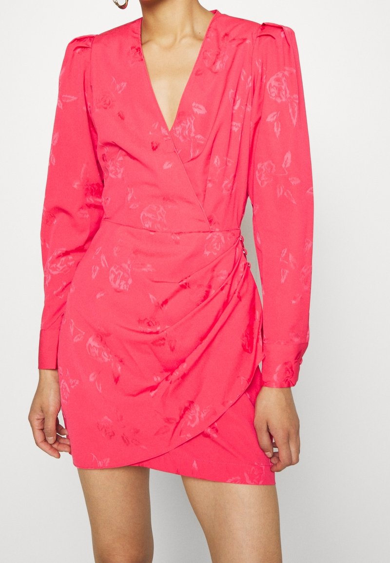 Femme portant une mini-robe rose vif à manches longues avec des motifs floraux subtils et un devant cache-cœur avec des boutons latéraux.