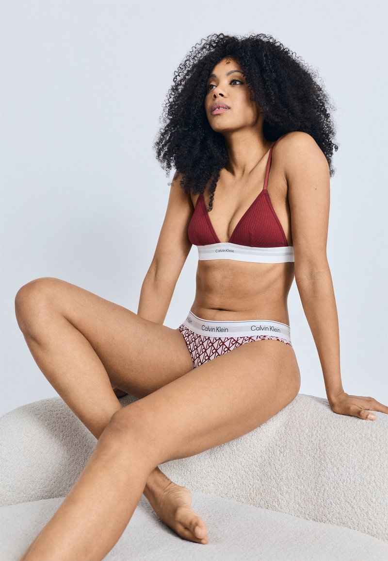 Bordový rebrovaný trojuholníkový bralette a vzorované červeno-biele bikiny. Mäkká látka s elastickými pásmi s logom. Modelka sediaca na textúrovanom povrchu.