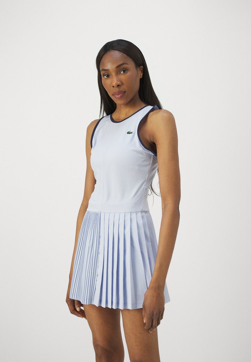 Hellblaues Kleid mit einem taillierten Oberteil und einem plissierten Rock, aus glattem Stoff gefertigt. Verfügt über einen marineblauen Saum und ein kleines grünes Logo auf der Brust.