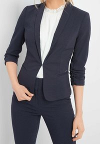 Granatowa marynarka w delikatny wzór w kropki, o dopasowanym kroju, zapinana na jeden guzik, z rękawami do połowy przedramienia, zestawiona z białą bluzką.