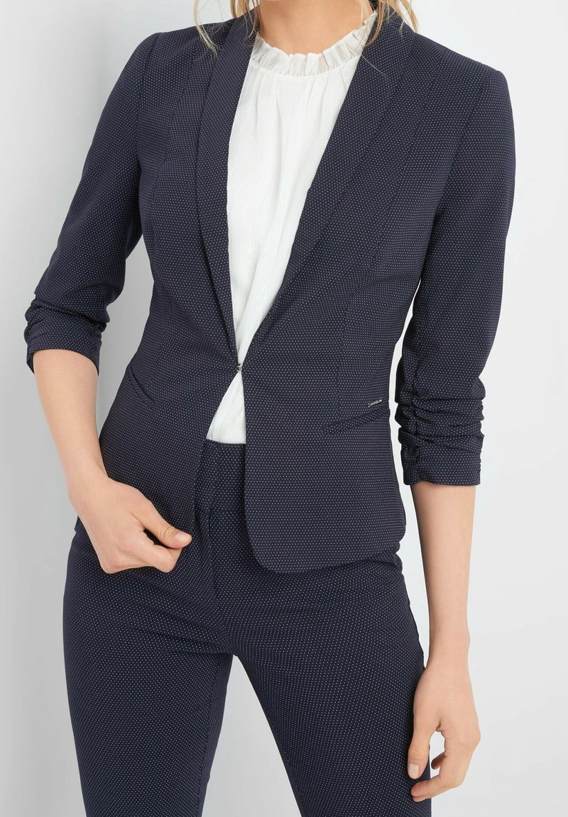 Granatowa marynarka w delikatny wzór w kropki, o dopasowanym kroju, zapinana na jeden guzik, z rękawami do połowy przedramienia, zestawiona z białą bluzką.