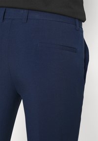 Närbild av en person som bär skräddarsydda marinblå byxor med passpo pocket och bälteshällor, tillsammans med en svart topp.