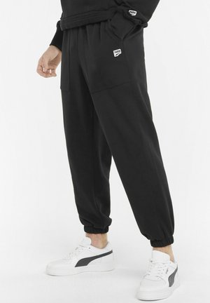 Pantalones jogger negros con un corte relajado, tobillos elásticos, dos bolsillos delanteros y un pequeño parche con el logo. Combinados con zapatillas blancas.