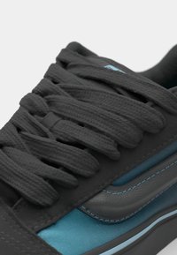 Zwarte sneaker met gestructureerde donkergrijze veters, blauwe accenten en een glad paneel dat lijkt op leer aan de zijkant. Subtiele stiksels zichtbaar.