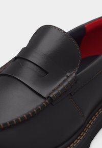 Μαύρο δερμάτινο παπούτσι τύπου penny loafer με λεπτομερή καφέ ραφές και κόκκινη εσωτερική επένδυση, εμφανιζόμενο σε κοντινή λήψη.