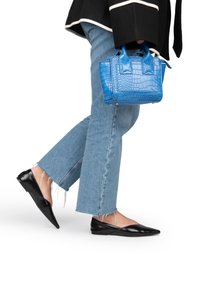 Blauwe handtas met krokodillenprint en twee bovenhandvatten, gecombineerd met zwarte puntige platte schoenen en lichtblauwe jeans met rafelige zoom.