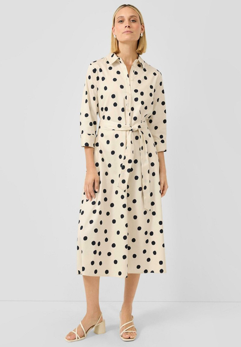 Kobieta ubrana w kremową midi koszulową sukienkę w czarne groszki, z rękawami 3/4, wiązaniem w talii oraz beżowymi sandałami na obcasie.