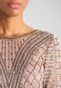 Pärlad klänning med en naken bas och intrikata mönster, inklusive paljetter och metalliska accenter på halsen och ärmarna.