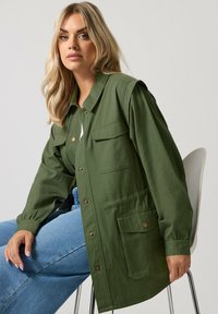 Vaalea nainen istuu tuolilla yllään suuri vihreä nappijakku, jossa on taskut, ja vaaleansiniset farkut yksinkertaisen taustan edessä.