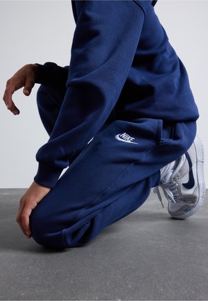 Dunkelblauer Nike-Sweatshirt und Jogginghose aus weichem Material. Weißes Nike-Logo auf der Hose. Die Schuhe sind weiß mit blauen Akzenten und grauen Sohlen.