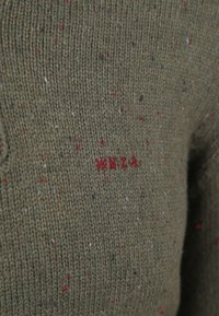 Groene gebreide trui met een gestructureerd oppervlak, voorzien van kleurrijke spikkels in rood, blauw en wit, en een geborduurd "NZ" logo.